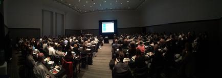 A large conference room with rows of people sitting in chairs facing a stage. The room has a high ceiling with recessed lighting and a large screen at the front of the room displaying a presentation. The people in the room appear to be engaged in a discussion or presentation with some people standing at the podium and others sitting at tables. The audience is attentively listening to the presentation.