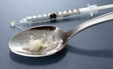 A silver spoon and a syringe next to it. They are resting on a blue grey surface. On the spoon there is a small amount of white substance likely an opioid or a another drug. The substance is spilling out of the spoon and onto the tip of the syringe. 