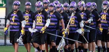 A group of lacrosse players walking on the field. They are all wearing purple and yellow jerseys with the number 66 on the front and the number 44 on the back. The players are also wearing helmets and holding lacrosse sticks. The field is covered in grass and there is a fence in the background. The image appears to have been taken during a game.