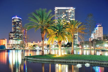 A night view of a city skyline with tall buildings and skyscrapers. The sky is dark blue and the buildings are lit up with colorful lights. In the foreground there is a body of water with three palm trees in the middle. The water is calm and reflects the lights from the buildings. On the right side of the image there are several white swans swimming in the water. The buildings in the background are tall and modern with a mix of high-rise and low-rise structures. The overall atmosphere is peaceful and serene.