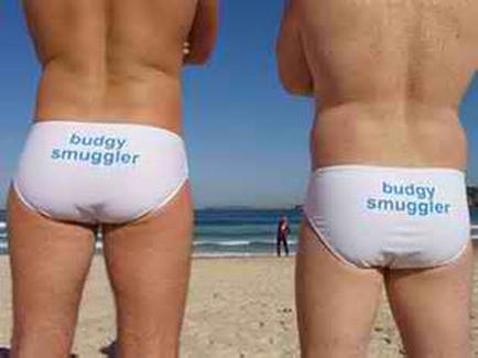Two men standing on a beach with their backs to the camera. They are both wearing white underwear with the words "buddy smuggler" written on them in blue. The man on the left is standing with his back to the viewer while the man in the middle is standing behind him. The background shows the ocean and a clear blue sky. The men appear to be in their late twenties or early thirties.