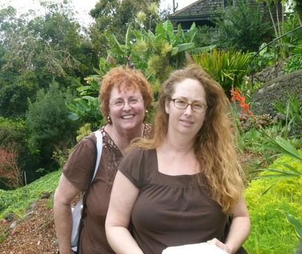 Two women standing side by side in a garden. They are both smiling and appear to be happy. The woman on the left is older and has red hair while the woman in the middle is younger. Both women are wearing brown tops and glasses. The garden is filled with various plants and trees and there is a stone wall in the background. The sky is overcast and the overall mood of the image is cheerful and relaxed.