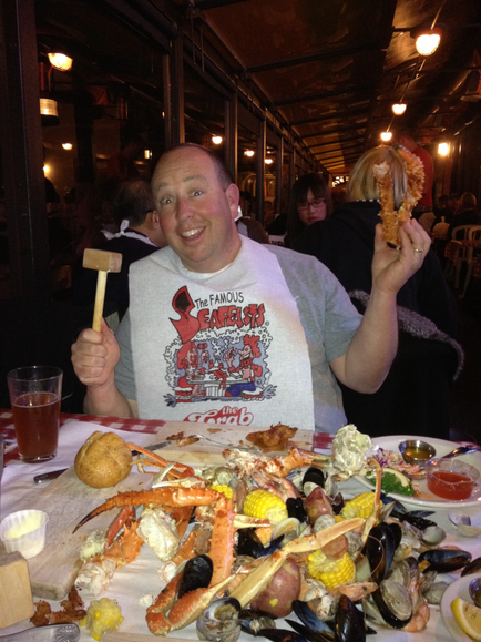 A man sitting at a table in a restaurant. He is wearing a grey t-shirt with a red graphic on it that reads "The Famous seafood". He is holding a wooden mallet in his right hand and is smiling at the camera. On the table in front of him there is a large platter of seafood including clams mussels corn and crab legs. There is also a glass of beer on the table. The restaurant has a high ceiling with wooden beams and there are other people in the background.