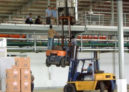A large warehouse with a forklift truck in the foreground. The forklift is orange and blue in color and has the word "TOYOTA" written on it. There are several workers in the background some standing and some sitting on the floor. The warehouse has a high ceiling with metal beams and a conveyor belt running along it. On the left side of the image there are several stacks of cardboard boxes. The workers appear to be working in the warehouse.