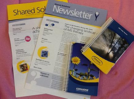 Three items on a pink surface. On the left side there is a yellow folder with the words "Shared Solutions Newsletter" written on it. Next to it there are two pages of a newsletter with a blue cover and white text. The newsletter is titled "The changing landscape of MS therapy" and has an image of a sunflower on the cover. The notebook on the right side has a blue and yellow cover with the word "Copaxone" written in white text and a picture of a tree. The pages appear to be new and unused.