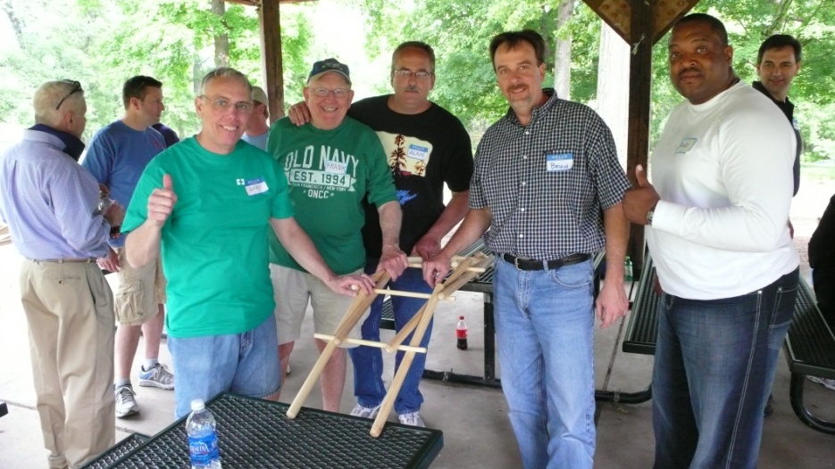 A group of five men standing around a table in a covered area with trees in the background. They are all smiling and five of them appear to be posing for a photo. The man in the center is holding a wooden structure which appears to be a ladder. In the background there are other people standing around not looking at the camera. There is a water bottle on the table.