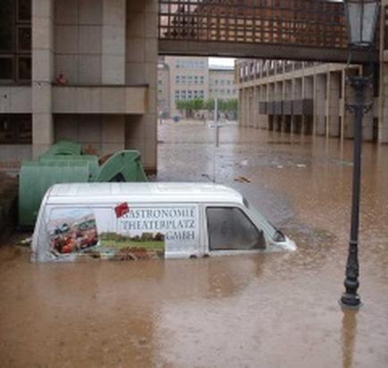 A flooded street with a white van partially submerged in the water. The van has a banner on the side that reads "astronomic Theaterplatz GmbH". The street is lined with buildings and there is a street lamp on the right side of the image. The water is up to the level of the van and there are debris scattered around. The sky is grey and cloudy indicating that it is raining.