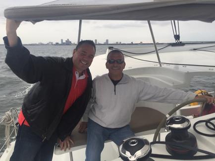 Two men on a sailboat in the ocean. The man on the left is wearing a black jacket and a red shirt and he is holding up a gray sail with both hands. He is smiling and appears to be happy. The other man is sitting at the helm of the boat wearing a gray sweatshirt and sunglasses. In the background there is a city skyline visible on the horizon. The water is calm and the sky is overcast.