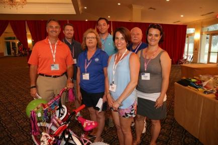 A group of seven people standing in a room with a red curtain in the background. They are all smiling and appear to be posing for a photo. On the left side of the image there is a man wearing an orange polo shirt and khaki pants. Next to him there are two women wearing blue shirts and shorts one wearing a blue shirt and the other wearing a gray tank top. In the center of the group is a woman wearing a pink tank top and shorts. She is holding a pink and white bicycle with colorful decorations on the front. Behind them is a table with various items on it. The room appears to be a conference or event space with a chandelier hanging from the ceiling.