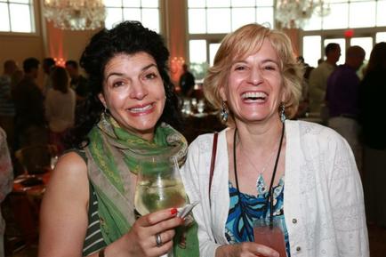 Two women standing in a large room with large windows and chandeliers hanging from the ceiling. They are both smiling and holding wine glasses in their hands. The woman on the left is wearing a green scarf and has dark hair while the woman in the middle has blonde hair. Both women appear to be happy and enjoying themselves. In the background there are other people in the room suggesting that they are at an event or gathering.
