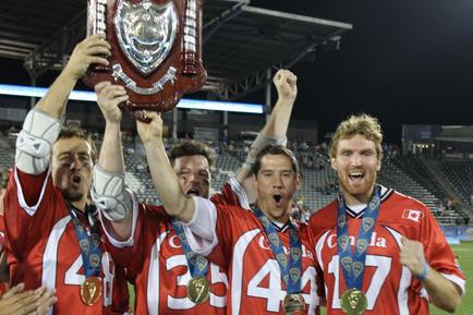 A group of four soccer players from the same team all wearing red jerseys with gold medals around their necks. They are standing on a field with a stadium in the background. The player in the center is holding up a large trophy with a shield on top which appears to be the ufa Champions League trophy. The players are smiling and cheering with their arms raised in triumph. The image appears to have been taken at night as the stadium is lit up with floodlights.