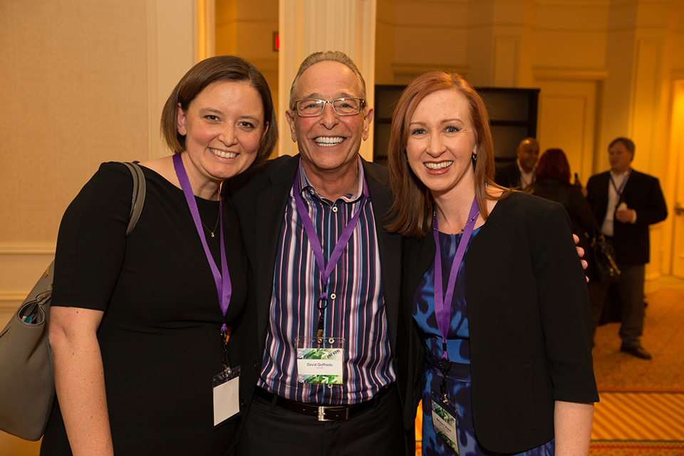 Three people standing in a large room with a high ceiling. The person in the middle is an older man with glasses and a striped shirt. He is smiling and posing for the photo with two women on either side of him. All three are wearing purple lanyards around their necks. The woman on the left is wearing a black dress and carrying a gray purse. The woman on the right is also wearing a blue dress and a black blazer. They are all smiling and appear to be happy. In the background there are other people in the room and a carpeted floor.