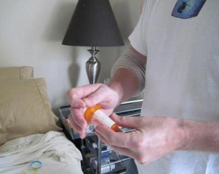 In this image we can see a person's hands holding an orange-colored pill bottle. The person is wearing a white t-shirt with a blue logo on it. In the background there is a bed with a beige blanket and a lamp on a nightstand. The bed appears to be in a bedroom.