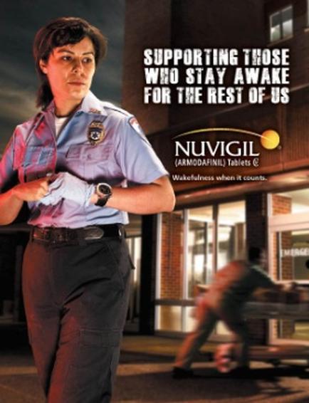 A promotional poster for vigil (Armodafinil) tablets. It features a woman in a police uniform walking on a street at night. She is wearing a blue uniform with a badge on the left side of her chest and a belt with a buckle. The woman has shoulder-length dark hair and is looking off to the side with a serious expression on her face. In the background there is a building with a sign that reads "Wakefulness when it counts." On the right side of the image there are two people running away from the building. The text on the image reads "Supporting those who stay awake for the rest of us."
