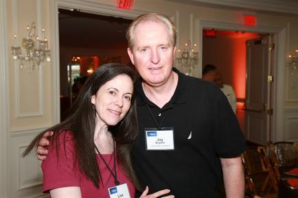 A man and a woman standing in a room with chandeliers and other people in the background. The man is on the right side of the image and the woman on the left side is standing close to him. They are both smiling and appear to be posing for the photo. The woman is wearing a red t-shirt and has long dark hair while the man has short blonde hair. Both of them are wearing name tags around their necks. The room appears to be a formal event space with a chandelier hanging from the ceiling and a door leading to another room.