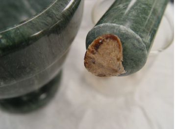 A close-up of a green mortar and pestle. The pestle is on the right side of the image and appears to be resting on a clear plastic cup. The pestle has a brown gummy looking object stuck to it possibly a crushed pill. The background is blurred but it seems to be a white surface.