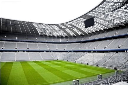 The interior of a large sports stadium. The stadium has a curved roof with a skylight above it allowing natural light to enter the space. The seats are arranged in rows facing the field. The field is covered in artificial turf and there is a goalpost on the right side of the image. The sky is visible through the skylights and the overall atmosphere of the stadium is bright and airy.
