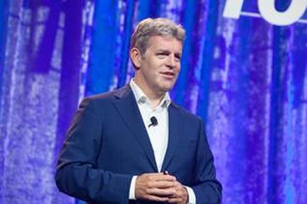 A man standing on a stage with a blue curtain in the background. He is wearing a dark blue suit and a white shirt. He has short grey hair and is holding a microphone in his left hand. He appears to be giving a presentation or giving a speech. The text on the curtain behind him reads "70".