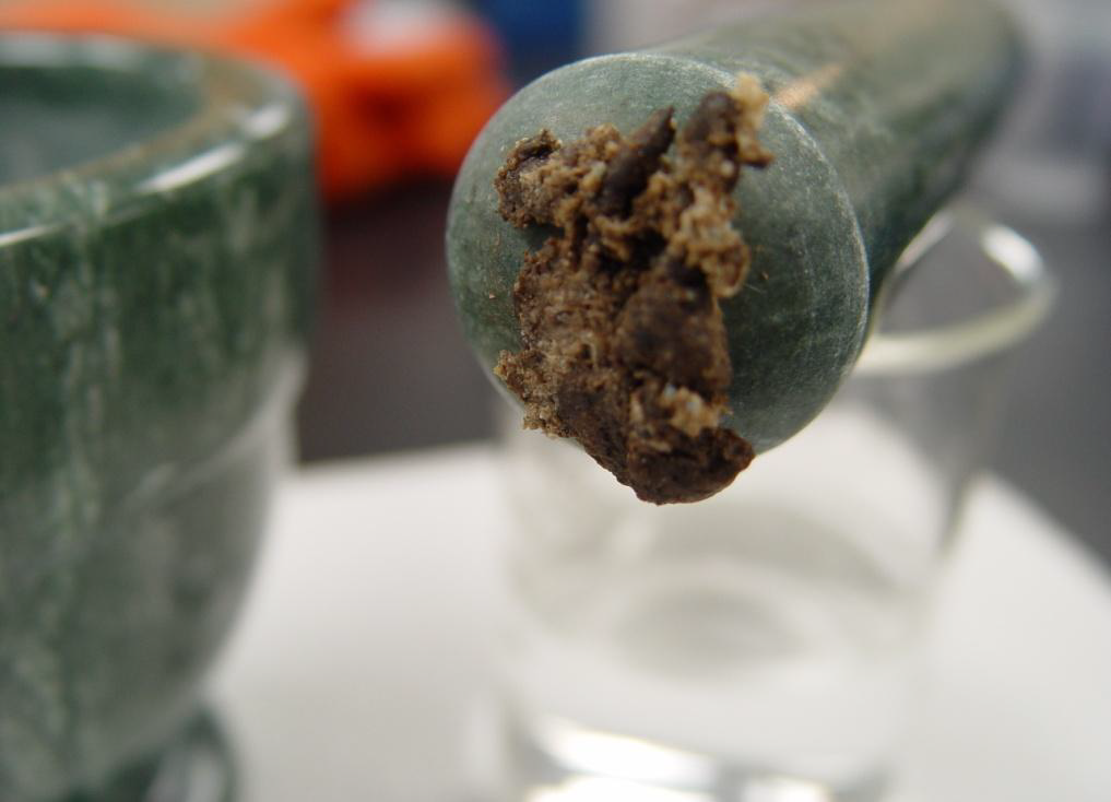 A close-up of a green mortar and pestle. The pestle is on the right side of the image and appears to be resting on a clear plastic cup. The pestle has a brown powdery looking substance stuck to it possibly a crushed pill. The background is blurred but it appears to be a white table or countertop at the center of a room.