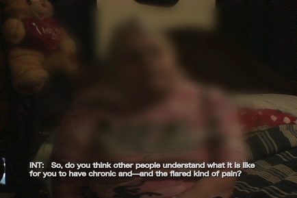 A person lying on a bed with a teddy bear on the left side of the bed. The person is wearing a pink shirt and appears to be in a state of distress. The background is blurred but it seems like the person is in a bedroom. The text on the image reads "INT: So do you think other people understand what it is like for you to have chronic and-and-the flared kind of pain?"