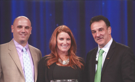 Three people standing on a stage with a blue curtain in the background. On the left is a bald man wearing a beige suit and tie on the right is a middle-aged woman with long red hair and a necklace and in the center is a man with short dark hair. All three are smiling and posing for the photo. The woman is holding a trophy which appears to be an award or a trophy. The man on the left has short dark hair and is wearing a suit and a green tie. The background is a solid blue color.