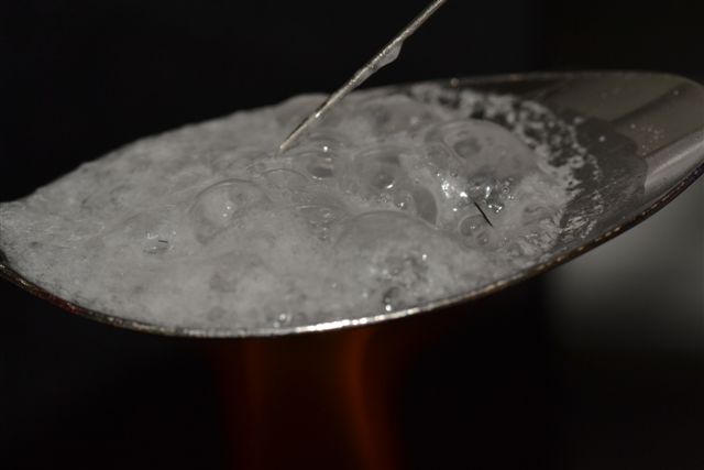 A close-up of a spoon with a white substance on it likely an opioid or another drug. The substance is bubbling as though it was just heated. A tip of a syringe is in the substance.  The background is dark making the spoon the focal point of the image.