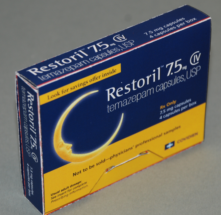 A photograph of a blue and yellow box of Restoril 7.5 mg capsules. The box is rectangular in shape and has a white background. The front of the box has the brand name "Restoril" written in bold white letters at the top followed by "7.5mg Capsules" in smaller white letters. Below that there is an image of a crescent moon with the text "Look for savings offer inside" and "Temazepam capsules USP" in yellow letters. <br /><br />The box also has a warning label that reads "Not to be sold-physicians' professional samples" in black letters. The label also mentions that the capsules are not available for sale. There is also a small illustration of a syringe on the bottom right corner of the label.