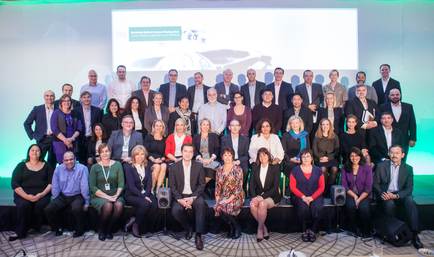 A large group of people posing for a group photo on a stage. There are around 20 people in the photo all dressed in formal attire. They are arranged in a semi-circle facing the camera and smiling at the camera. The background of the photo is a large screen with a green and white design and there are speakers on either side of the screen. The people are of different ages genders and ethnicity's and they appear to be of different ethnicity's. Some of them are wearing suits and ties while others are wearing casual clothing. The overall mood of the image is happy and celebratory.