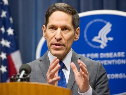 A man standing behind a podium with an American flag in the background. He is wearing a suit and tie and appears to be speaking into a microphone. Behind him there is a blue banner with the logo of the United States Air Force and the words "For Disease Prevention" written in white. The man is gesturing with his hands as he speaks. He looks serious and focused.