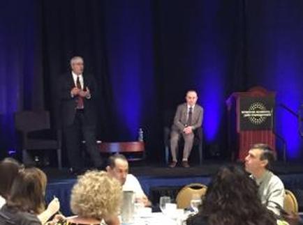 A stage with a blue curtain and a podium on the right side. On the left side of the stage there is a man standing and speaking into a microphone. He is wearing a suit and tie and appears to be giving a presentation. In front of him there are two men sitting on chairs facing each other. The man on the left is sitting on a chair and is speaking into the microphone. The other man is sitting next to him and is listening attentively. There are people sitting at tables in front of them some of them are engaged in conversation. The audience is seated in chairs and some are looking at the man speaking.