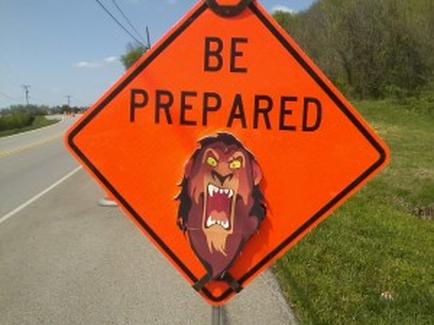 Of a triangular orange sign with the words "BE PREPARED" written in bold black letters. The sign is attached to a metal pole on the side of a road. In the center of the sign there is an illustration of a lion's head with its mouth open showing its sharp teeth. The lion's fur is a deep brown color and its eyes are a bright yellow. The background of the image shows a grassy field and a blue sky.