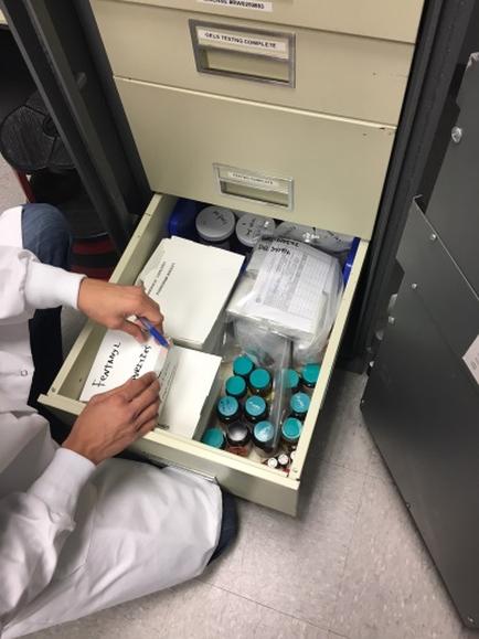 A person wearing a white lab coat sitting on the floor in front of a filing cabinet. The filing cabinet is open and the person is holding a pen and writing on a piece of paper. Inside the filing cabinet there are several small bottles of blue-colored liquid a white box and a few other items. The person appears to be working on a project or organizing the contents of the cabinet.