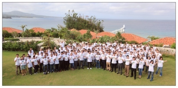 A large group of people standing on a grassy hill overlooking the ocean. They are all wearing white t-shirts. In the background there are houses with red roofs and a view of the ocean and mountains. The sky is blue and the weather appears to be sunny and warm. The group seems to be posing for a group photo.