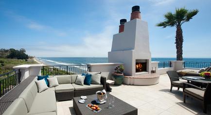 A beautiful outdoor patio area with a stunning view of the ocean. The patio is furnished with a large sectional sofa with blue and gray throw pillows a coffee table with a tray of fruit and a vase of flowers and several chairs and tables. There is also a fireplace with a white chimney and a palm tree on the right side of the patio. The ocean can be seen in the background with a clear blue sky above. The overall atmosphere of the space is peaceful and relaxing.