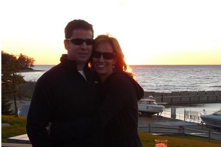 A man and a woman standing on a balcony overlooking the ocean at sunset. The man is on the left side of the image wearing a black jacket and sunglasses and the woman on the right side is wearing a gray jacket and black sunglasses. They are both smiling and looking at the camera. In the background there are several boats docked on the water and a small island with trees on the shore. The sky is orange and pink indicating that it is either sunrise or sunset.