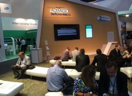 A booth at an international trade show. The booth is called "adagio" and has a large screen on the wall displaying information about the company. There are several people sitting on white sofas in front of the booth engaged in conversation. On the left side of the image there is a sign that reads "International Booth". The booth appears to be well-lit with natural light coming in from the windows.