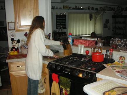 A young woman standing in a kitchen next to a gas stove. She is wearing a white sweater and blue jeans and is holding a knife in her hand. The kitchen has wooden cabinets and a countertype with various kitchen items on it. On the countertype there is a red kettle a bottle of condiments and a few other kitchen utensils. There is also a Mickey Mouse figurine on the kitchen counter. The woman appears to be preparing food or preparing a meal.