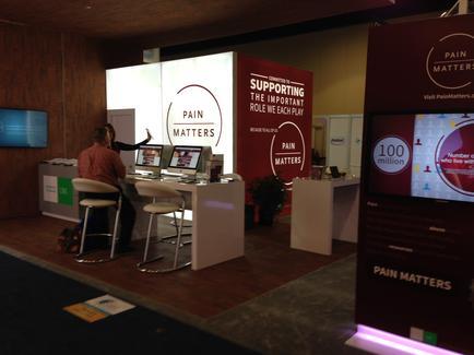 A trade show booth with a red and white color scheme. The booth has a large screen on the right side with the words "Pain Matters" written in bold white letters. On the left side of the booth there is a desk with two computer monitors and a person sitting at a desk in front of it. The person is wearing a red shirt and appears to be working on one of the monitors. In the background there are other booths and a large TV screen. The overall atmosphere of the image is professional and modern.