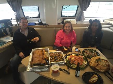 Three people sitting at a table in a restaurant. The table is covered with a variety of food items including sandwiches salads and drinks. The person on the left is holding a plate of food while the person in the middle is wearing a pink sweater. The two women on the right are wearing black jackets. They are all smiling and appear to be enjoying their meal. The restaurant has large windows with curtains and there is a desk in the background.