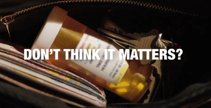 A close-up of a black purse with a zipper open. Inside the purse there is a small orange prescription bottle with a white label that reads "Don't think it matters?" The bottle is lying on top of a stack of magazines. There are also a few keys scattered around the purse. The background is dark and out of focus.