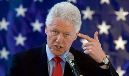 Former US President Bill Clinton speaking into a microphone. He is wearing a dark suit and a red tie. The background is blurred but it appears to be an American flag. Clinton is gesturing with his right hand as he speaks. He has a serious expression on his face and is looking directly at the camera.