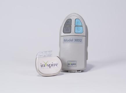 A model 3032 which is a device used to measure the temperature of a person's body. The device is grey in color and has a rectangular shape with rounded edges. It has a control panel on the top with three buttons - a blue button a green button and a white button. On the left side of the device there is a small white container with the brand name "inspire" written on it. The container appears to be made of plastic and is placed next to the device. The background is plain white.