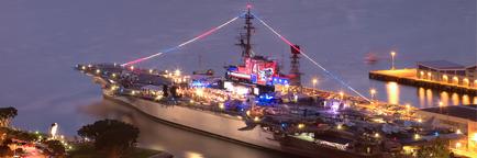 An aerial view of a large ship docked at a pier at night. The ship is illuminated with colorful lights creating a vibrant and lively atmosphere. The deck of the ship is filled with people and vehicles and there are several antennas and other equipment visible on the deck. The sky is dark indicating that it is nighttime. The pier is lined with buildings and trees and the water is calm and still. The overall mood of the image is lively and festive.