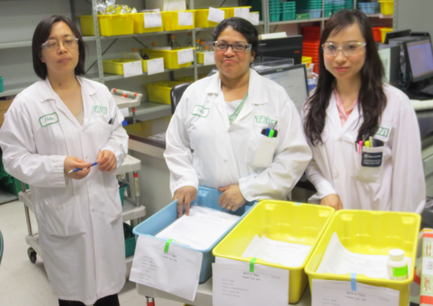 Three women in a laboratory setting. They are all wearing white lab coats with green labels and are standing in front of a table with three yellow plastic bins. The woman on the left is holding a blue tray with a piece of paper in it while the woman in the middle is holding an object in her hand. All three women are smiling and appear to be posing for the photo. In the background there are shelves with various laboratory equipment and supplies.