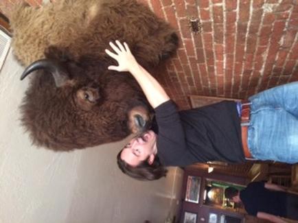 A young man standing in front of a large stuffed bison. The bison is lying on its side on the floor with its head resting on the man's shoulder. The man is wearing a black t-shirt blue jeans and a red belt. He is reaching up to touch the bison's head with his right hand. The background appears to be a brick wall with a window on the left side.