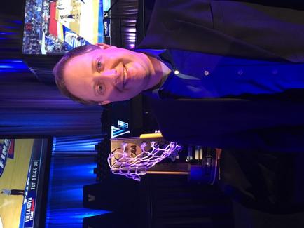 A man standing in front of a basketball hoop. He is wearing a dark blue suit and is smiling at the camera. The hoop is made of metal and has a net attached to it. In the background there is a large screen displaying a basketball game. The room is dimly lit with blue lights.