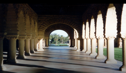 A long arched corridor with columns on either side. The corridor is made of stone and has a tiled floor. The columns are tall and slender with intricate carvings on the top and sides. The archway is supported by a stone wall on the left side and the columns are arranged in a symmetrical pattern. In the background there are palm trees and a blue sky. The image is taken from a low angle looking up at the corridor. The overall mood of the image is peaceful and serene.