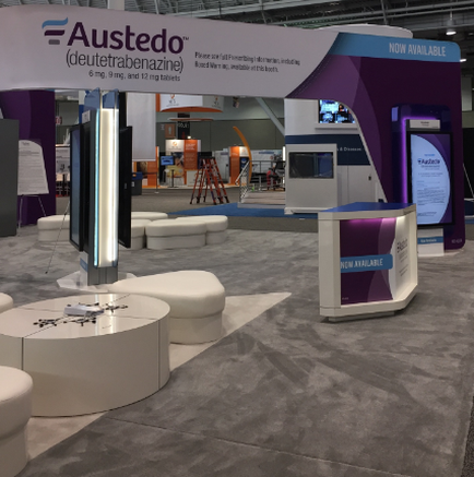 A trade show booth with a purple and white color scheme. The booth has a large banner that reads "busted (deutetrabenazine) - Now Available" in bold white letters. There are two large screens on the right side of the booth one on the left side and a smaller screen on the top right corner. In front of the screens there is a white reception desk with a blue countertype and a sign that says "Now Available". The booth is surrounded by white round tables and chairs and there are other booths and equipment in the background. The floor is made of gray tiles and the overall atmosphere of the trade show is modern and professional.