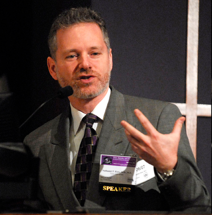 A man sitting at a podium with a microphone in front of him. He is wearing a gray suit and a purple tie. He has a name tag that reads "SPEAKER" and is gesturing with his right hand as if he is speaking. The background is dark and there is a window behind him. The man appears to be giving a presentation or giving a speech.
