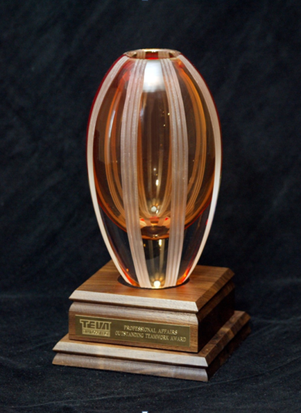 Of a trophy or award. It is a tall cylindrical object with a pointed top and a flat base. The body of the object is made up of multiple layers of glass with the top layer being orange and the bottom layer being white. The glass has a striped pattern with vertical lines running horizontally across it. The base is made of a dark wood with a gold-colored plaque on the front that reads "tea Professional Award." The background is black making the trophy stand out.